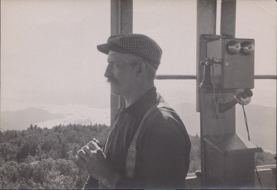 Image of an unidentified Forest Ranger inside the Black Mountain fire tower. The Ranger holds a pair of binoculars as he peers into the distance. Lake George is visible in the background.