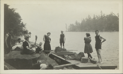 Photograph of 7 men and three women wearing bathing suits and waving at the camera with rocks while riprapping an island on Lake George with John S. Apperson Jr.'s barge "Article 7 Section 7," c. 1910.