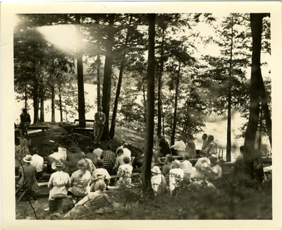Crowd of attendees listening to John S. Apperson Jr. and one other man lecturing in front of maps of the Adirondacks at his camp Chilhowie on Lake George, NY at a Forest Preserve Association of New York State meeting during the summer of 1938.