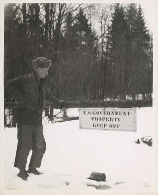Ed Richard standing next to a sign blocking the road reading "U.S. GOVERNMENT PROPERTY KEEP OFF" at the Panther Mountain Road at the boundary of Forest Preserve Land, in winter. This is a picture from a trip Paul Schaefer and the Adirondack Moose River Committee took to record illegal tree cutting activity at the site of the Panther Mountain Dam, circa 1946.
