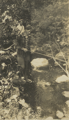 Paul Schaefer and an unidentified man, possibly his brother Vincent, fishing in a stream, circa 1930. Paul stands on a rock in the stream holding a fishing rod and a fishing basket wearing a plaid hunting jacket. The unidentified man is standing behind him wearing a white shirt and a newsboy hat.