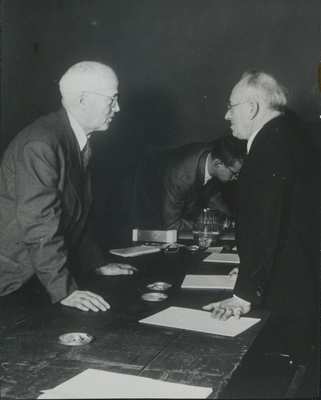 Photograph of John S. Apperson, Jr. leaning over a table to speak to Chauncey Hammond, a member of the Senate Judiciary Committee of New York State, during the two day Ostrander Amendment public hearings at the American Museum of Natural History in New York City on January 20-21, 1950.