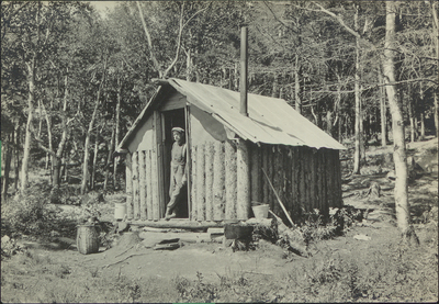 A man leaning against the doorway of an Adirondack a migrant hunting cabin, circa 1915.