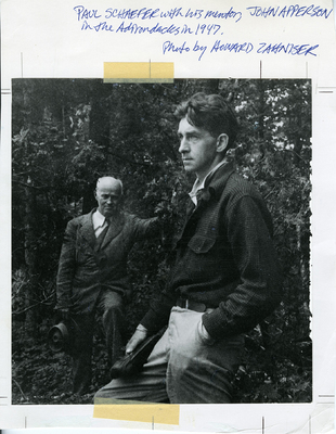 Photo of Paul Schaefer with his mentor John S. Apperson Jr. standing outdoors in the Adirondacks. Schaefer is dressed in a plaid button up shirt and looks off camera, while Apperson stands behind donning a suit and holding a hat. Photo taken by noted conservationists Howard Zanhiser.