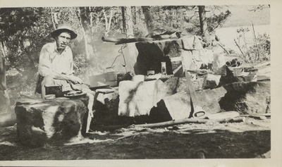 Man smiling and smoking a pipe while cooking over fire pit, surrounded by provisions and gear on Dollar Island, Lake George, NY, 1910. Called "a bum" by John S. Apperson, Jr. on verso. Additional information, possibly the figure's name, is noted by a later processor on verso in the 1990s.