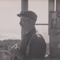 Image of an unidentified Forest Ranger inside the Black Mountain fire tower. The Ranger holds a pair of binoculars as he peers into the distance. Lake George is visible in the background.