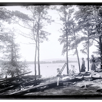 Two figures pose smiling over the remains of a dismantled squatter cabin on Big Burnt Island, Lake George, NY, 1917.