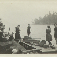 Photograph of 7 men and three women wearing bathing suits and waving at the camera with rocks while riprapping an island on Lake George with John S. Apperson Jr.'s barge "Article 7 Section 7," c. 1910.