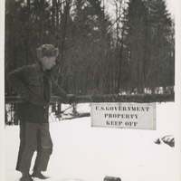 Ed Richard standing next to a sign blocking the road reading "U.S. GOVERNMENT PROPERTY KEEP OFF" at the Panther Mountain Road at the boundary of Forest Preserve Land, in winter. This is a picture from a trip Paul Schaefer and the Adirondack Moose River Committee took to record illegal tree cutting activity at the site of the Panther Mountain Dam, circa 1946.