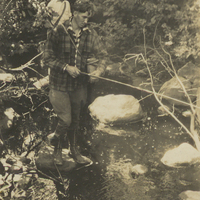 Paul Schaefer and an unidentified man, possibly his brother Vincent, fishing in a stream, circa 1930. Paul stands on a rock in the stream holding a fishing rod and a fishing basket wearing a plaid hunting jacket. The unidentified man is standing behind him wearing a white shirt and a newsboy hat.