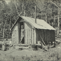 A man leaning against the doorway of an Adirondack a migrant hunting cabin, circa 1915.